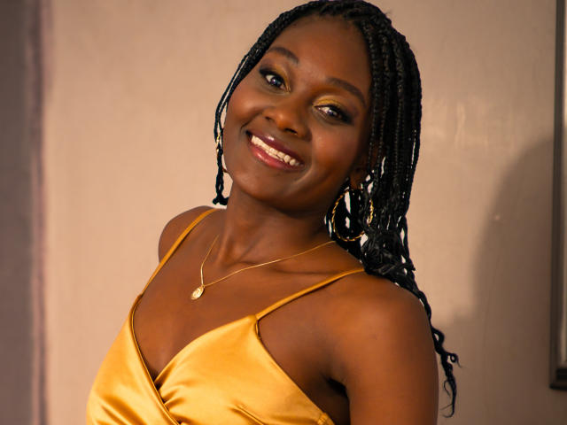 AndreaStorm with dark skin, smiling, wearing a yellow satin dress, gold necklace, and braided hair, showing her tits and a cute, sexy face.