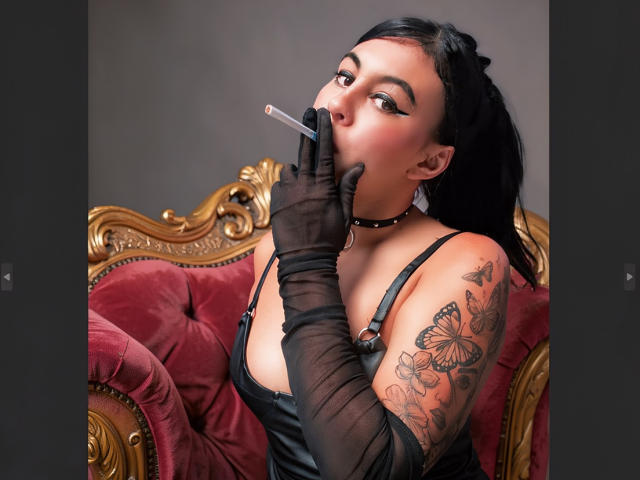 NenaBlack, con i capelli neri, indossa un collare nero e guanti trasparenti, mentre fuma una sigaretta, mostrando un braccio tatuato con una farfalla e un fiore, seduta su una poltrona di velluto rosso..