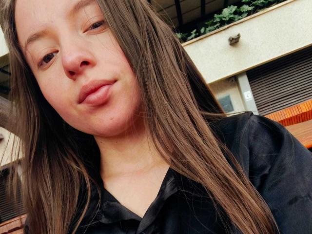 VioletaNova has puffy lips, long brown hair, and a slightly goofy expression, wearing a black shirt, with a small nose and light skin. She's a cute chick.