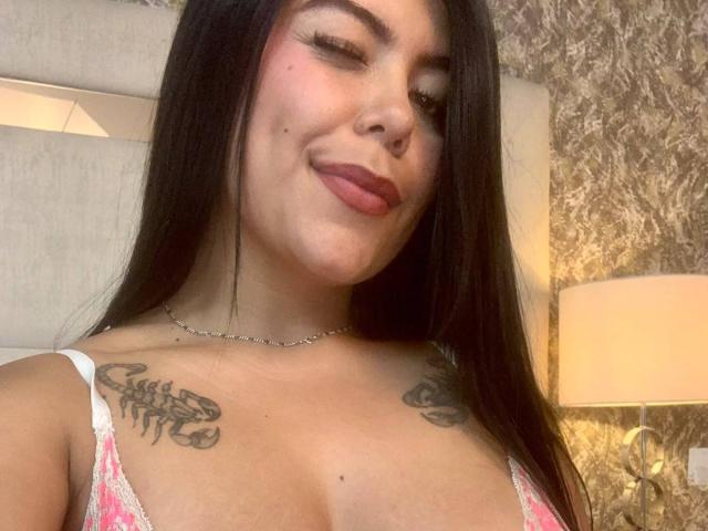 CatalinaReiyna with long black hair, slightly winking, pink lipstick, tattoos on her tits, wearing a pink and white lace top, necklace, and a subtle smile.