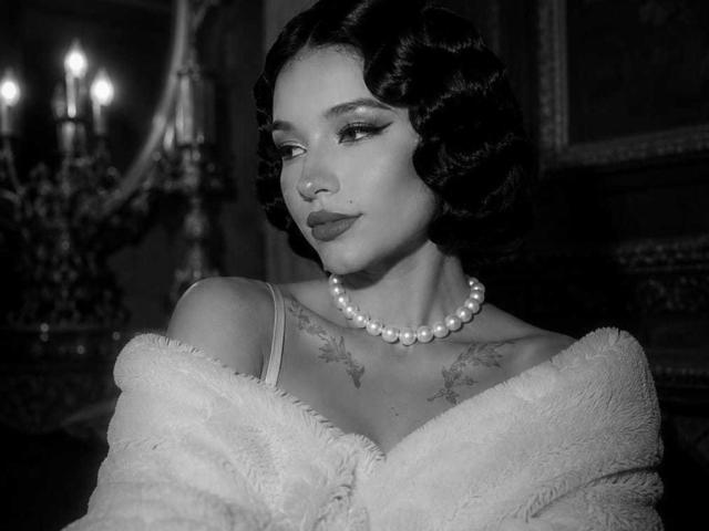 Photo en noir et blanc de CelesteOcampo, avec des cheveux courts et ondulés, foncés, un collier de perles et un manteau duveteux à épaules dénudées, montrant des tatouages sur sa poitrine, regardant vers le bas avec un sourire discret..