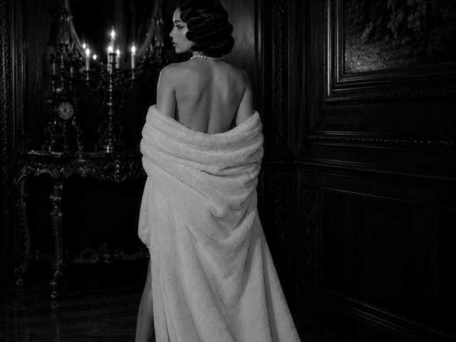 CelesteOcampo est dos à la caméra, avec une serviette blanche recouvrant ses seins et ses fesses, des cheveux noirs ondulés, un collier de perles, et se tient debout dans une pièce élégante, faiblement éclairée..