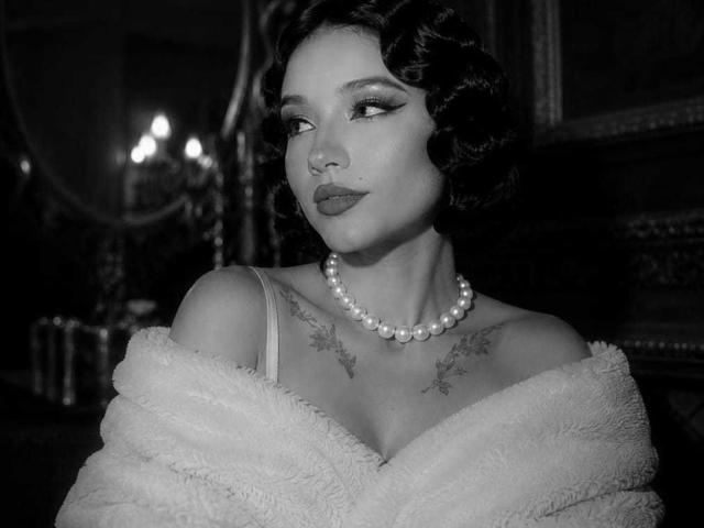 Fotografia in bianco e nero di CelesteOcampo con capelli scuri e ricci, collana di perle, vestaglia bianca soffice con spalline scoperte, che mostra tatuaggi sopra il seno, con un'espressione seducente e lo sguardo rivolto di lato..