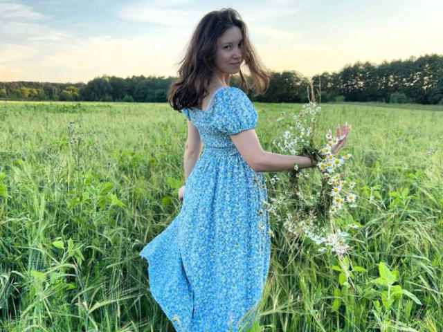 Kathrin Maddoy, vestida con un vestido azul con estampado floral, sosteniendo un ramo de flores silvestres, de pie en un campo alto de hierba verde, mirando hacia atrás con una ligera sonrisa, cabello castaño ondulado..