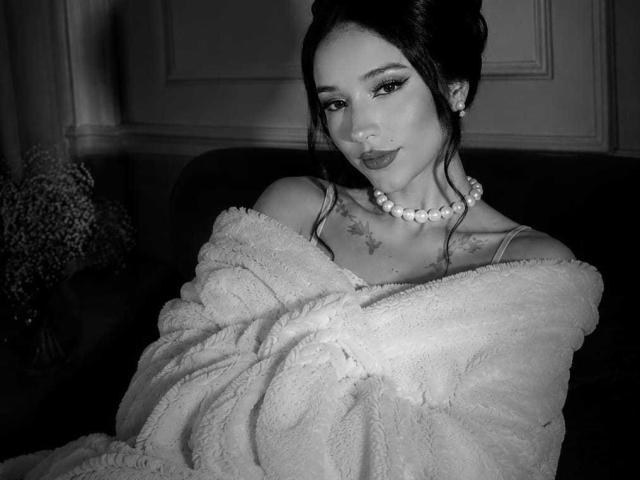 Celeste Ocampo, avec ses cheveux sombres attachés en chignon, portant une robe moelleuse à une épaule, un collier de perles et du maquillage, montrant des tatouages sur sa poitrine, regardant vers le bas avec un léger sourire..