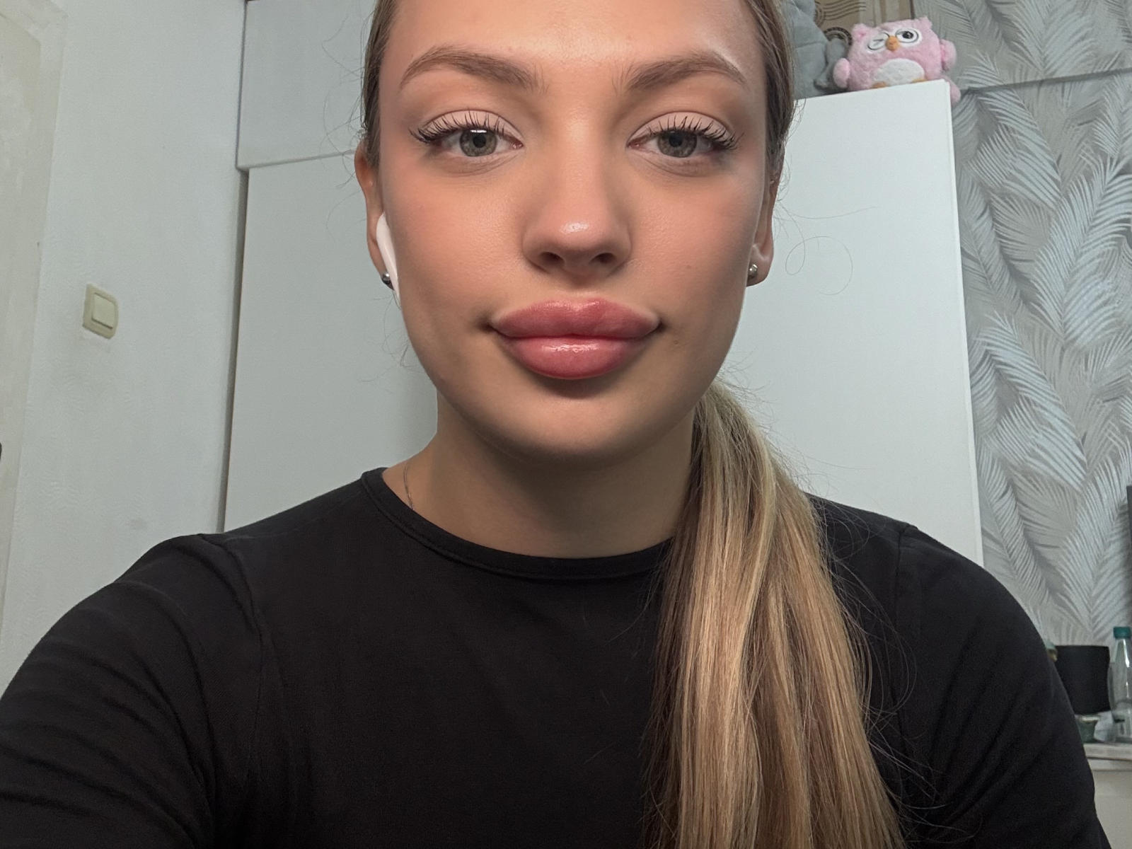DanyellFarhat, met een lichtbruine paardenstaart, een zwart shirt, roze glanzende lippen en witte oordopjes, die haar schattige achterwerk en kleine borsten laat zien in een selfie..