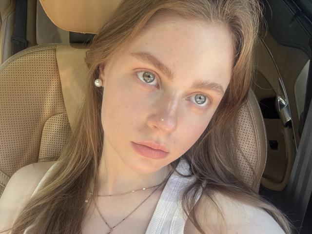 Virginia Eash, con ojos verdes, cabello castaño largo, piel pálida, vestida con una camiseta blanca y aretes de perlas, sentada en un coche con asientos beige y texturizados..
