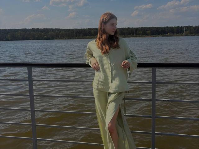 EdaAfan has pale skin, long brown hair, and is wearing a green outfit with a thigh slit, standing by a metal railing by a river, looking to the side.