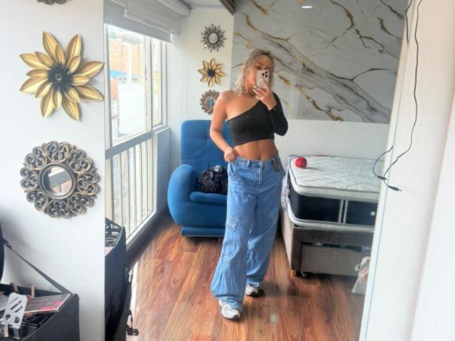 ElenaSaenz with blonde hair, black one-shoulder top, blue high-waisted jeans, and white sneakers, standing in a room, taking a mirror selfie, showing her ass.