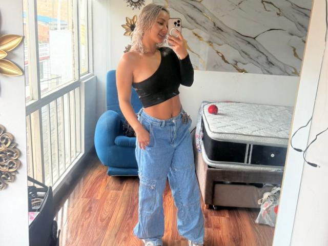 ElenaSaenz with blonde hair, black off-shoulder top, and blue cargo pants, standing in a room, taking a mirror selfie, showing her tits and ass, smiling.