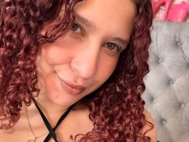 ValeryScot tiene el pelo rizado y rojizo, ojos verdes y un piercing en la nariz, sonríe con los labios ligeramente entreabiertos, lleva un top negro con tirantes y un collar dorado..
