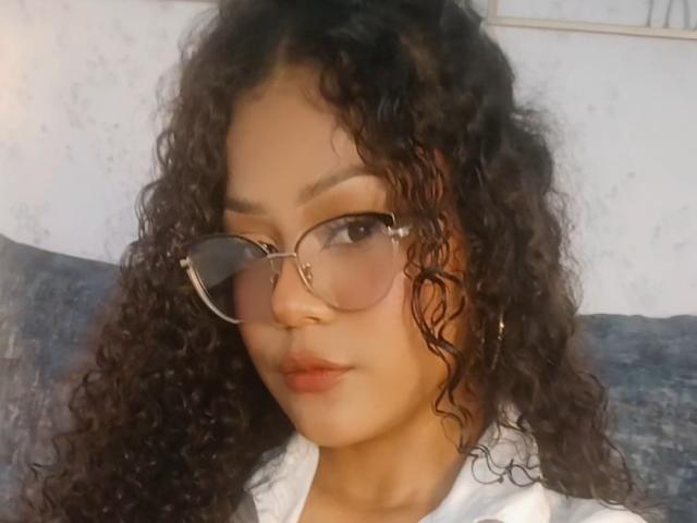 Zoely69 with curly black hair, wearing round glasses, and a white shirt, has a cute face and full lips, looking slightly to the side. Soft lighting.