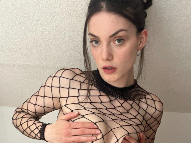 Nyrah-hot wearing a black fishnet top, hands covering her tits, pale skin, dark hair in a bun, green eyes, small hoop earrings, and a slightly open mouth.