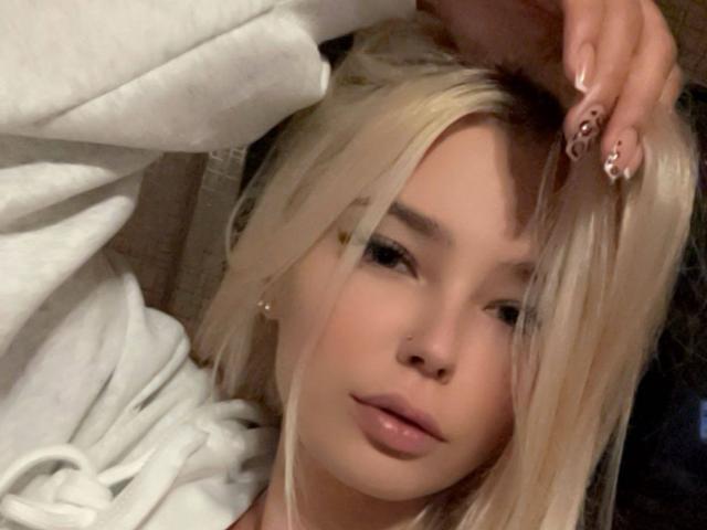 Adriene Degennaro ha i capelli biondi, un viso carino e indossa una felpa bianca. La sua mano, con unghie lunghe, rosa e scintillanti, tocca la sua testa. Trucco leggero e naturale..