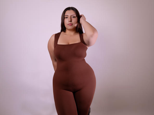 Sofie Malory, mit mittelbrauner Haut, langen, dunkelbraunen Haaren, trägt einen braunen, eng anliegenden, ärmellosen Body, steht, hat eine Hand im Haar, blickt in die Kamera, ist leicht kurvig mit großen, runden Brüsten..