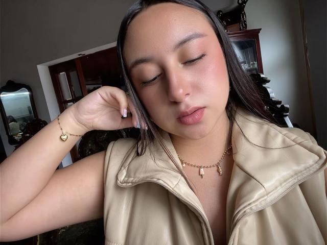 Rosalie Dupont, con los ojos cerrados, vistiendo un chaleco de cuero beige, un collar dorado y una pulsera, tocándose la cara, mostrando sus labios rosados y su cabello castaño liso..