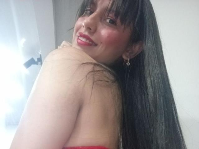 MadissonFontaine with long black hair, red lipstick, and earrings, smiling over her shoulder, showing her bare back and red strapless top, looking cute with a slight blush.