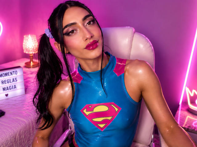 AlexaMissSx met een glanzende huid, zwarte vlechtjes, roze lippenstift, een blauw Supergirl kostuum, roze schouderbandjes, paars neonlicht en een witte stoel, zittend met zelfvertrouwen, kijkend naar de camera..