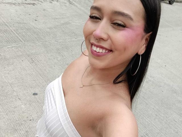 ZammySanz with pink blush on her cheeks, smiling, wearing hoop earrings, nose ring, white off-shoulder top, and a necklace, standing on a concrete surface.
