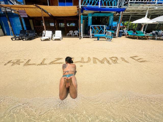 Victoria Waynes ajoelhada na areia molhada, de costas para a câmera, vestindo um biquíni azul de fio dental, com uma tatuagem no braço esquerdo, escrevendo "HALLOUMPHIE" na areia, praia, com estruturas azuis e de madeira ao fundo..