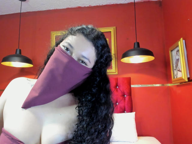Curly-haired AlanaNaur with fair skin, wearing a maroon mask, showing her large tits, red walls, black pendant lights, and a red tufted headboard in a red room.