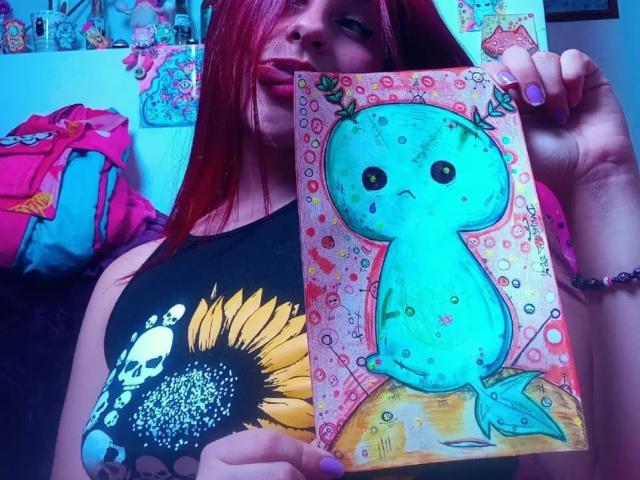 SophieHill with red hair holding a colorful, painted, blue-green, teardrop-eyed, bird-like creature drawing, wearing a black, sunflower, skull, and butterfly top, in a blue-lit room.