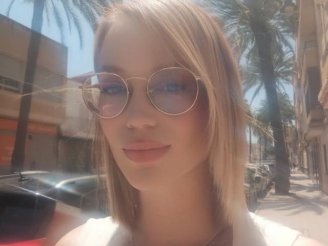 MissFrenchy, con un corte de pelo rubio, usando gafas de sol redondas, labios rosados y una blusa blanca, está de pie en una calle soleada con palmeras, mirando a la cámara, con reflejos de luz en las gafas..