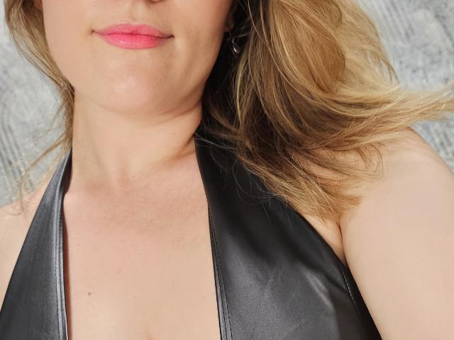 Le visage de Janet Jamesonn, avec ses lèvres roses, sa peau claire et ses cheveux blonds, porte un haut en cuir noir à encolure dégagée, laissant entrevoir une partie de sa poitrine et de sa clavicule. Ses seins sont partiellement visibles..