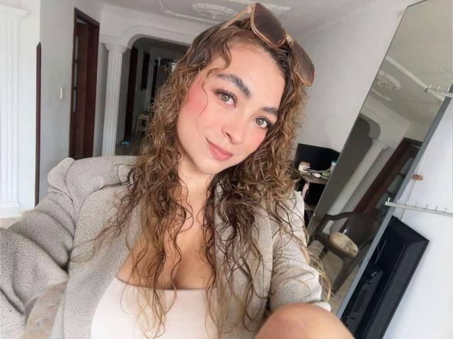 AriannaBlake with curly brown hair, pink blush on cheeks, gray cardigan, white top, small tits, subtle smile, sunglasses on head, taking selfie in bright room.