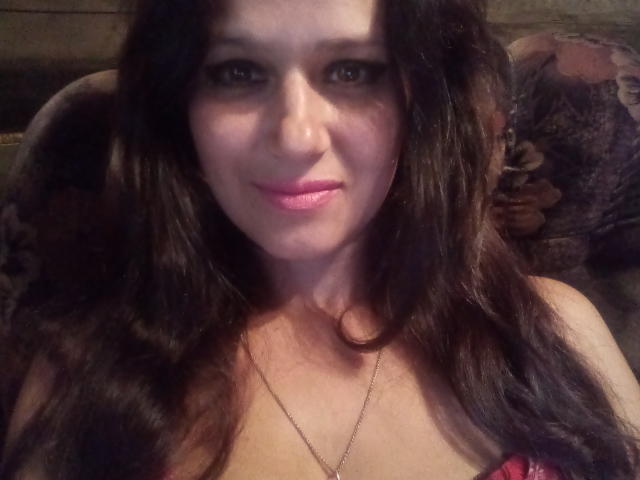 ZinaMillerx tiene el pelo largo, castaño oscuro, lápiz labial rosa y una ligera sonrisa. Lleva una blusa roja con un collar de oro delgado, mostrando sus pechos..