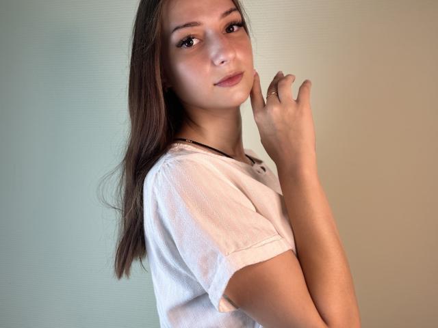 NoreenGriep with long brown hair, wearing a white tee, has a soft smile, raised hand with a ring, and a slight tan, looking at the camera.