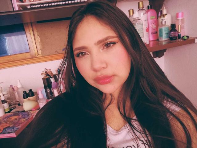 NicolDanniels has long brown hair, pink lips, and subtle eyeliner. She's wearing a white tank top, looking at the camera with a slight pout. Shelves with makeup are behind her.