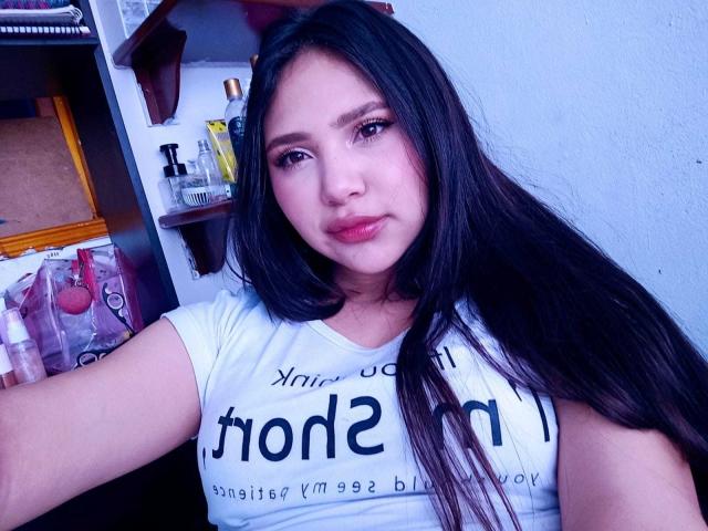 NicolDanniels has long black hair, light pink lipstick, and is wearing a white shirt. She's looking at the camera with a slight smile, showing her tits.