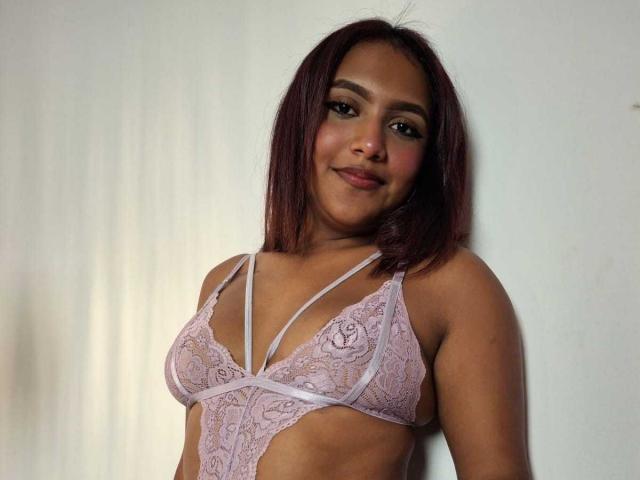 AmyMias with medium brown skin and red-brown hair, wearing a pink lace bra, showing her tits, smiling slightly, standing against a white wall.
