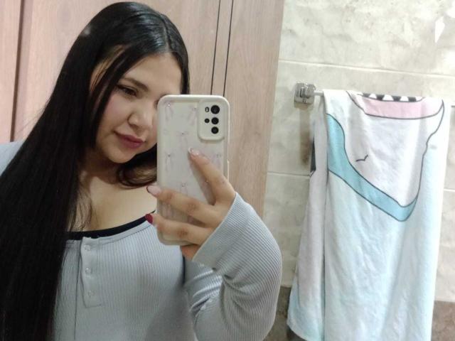NicolDanniels with long black hair, smiling, holding a phone, wearing a grey ribbed top, showing her small tits, in a bathroom, towel with pink and blue stripes.