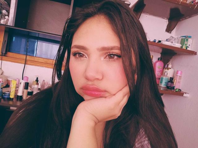 NicolDanniels has long brown hair, pouty pink lips, and her hand resting on her cheek with a serious expression. She's got thick eyelashes and a slightly pink face.