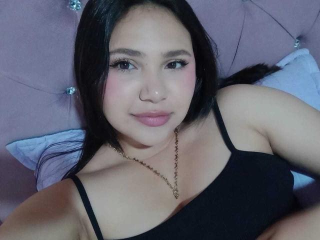 NicolDanniels has black hair, pink blush, and is wearing a black tank top with a gold necklace. Her tits are small, and she's smiling at the camera.