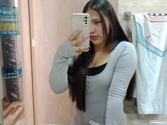 NicolDanniels with long black hair, wearing a grey long-sleeve top, taking a mirror selfie in a bathroom, showing her boobs slightly outlined under the top.