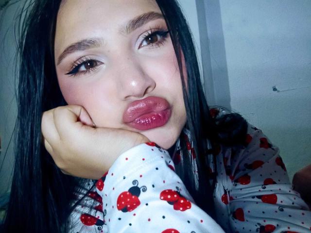 NicolDanniels has pouty red lips, long black hair, and is wearing a strawberry-patterned shirt. She's resting her chin on her hand, looking slightly to the side.