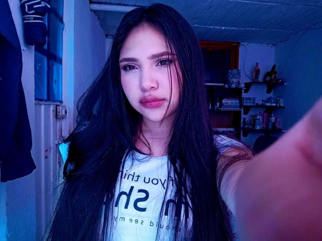 NicolDanniels with long black hair, blue eyes, and full lips, wearing a white shirt, taking a selfie in a blue-lit room. Her tits are small and her pussy is unseen.