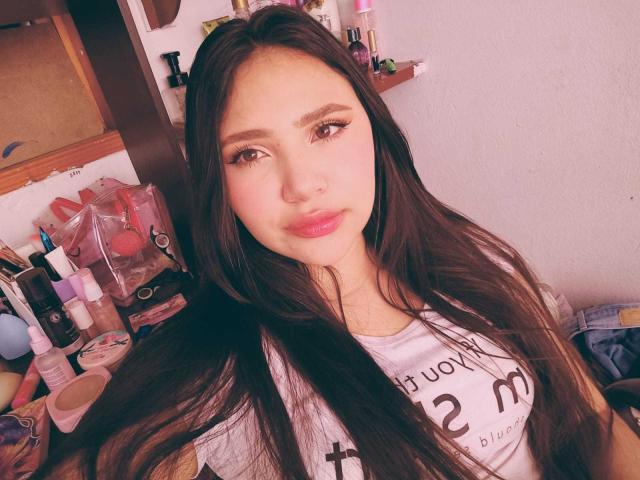 NicolDanniels has long brown hair, pink lipstick, and eyeliner, wearing a white shirt, sitting with makeup and toiletries in the background. Her tits are small, face is cute.