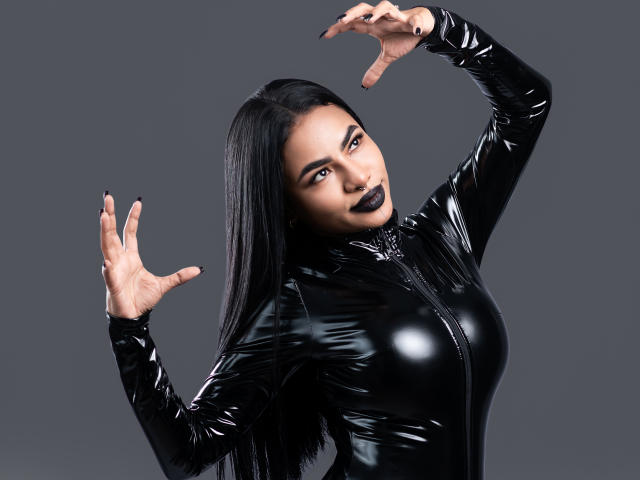 Anneroa, met lang zwart haar, draagt een glanzend zwart latex top, heeft haar handen omhoog, zwarte lipstick en een neusring, en kijkt met een lichte glimlach naar de camera..