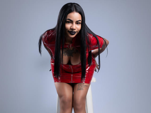 Anneroa mit langen, schwarzen Haaren, trägt ein glänzendes, rotes Latexgarderobe und beugt sich vor, wobei ihre Brüste sichtbar sind. Sie hat ein Tattoo auf der Brust und ein Tattoo an der Hüfte, trägt dunkelroten Lippenstift und steht vor einem Hocker..