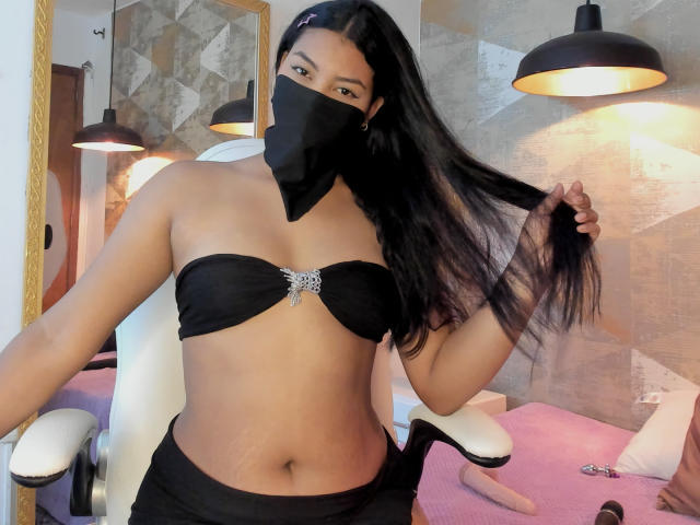 LalilaBrunette with dark hair, wearing a black mask and strapless black top with a gem, holding her hair, showing her flat stomach and black bottom, in a salon chair.