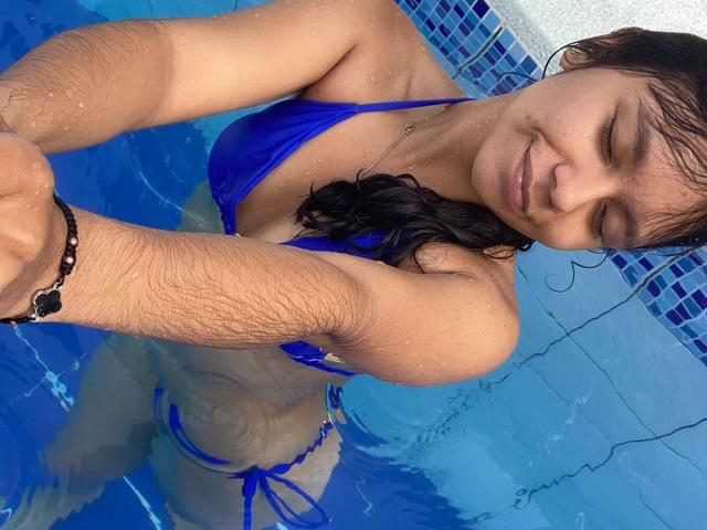 JuliaBennett with wet black hair, blue bikini, and tan skin, standing in a pool, smiling, showing her small tits and slightly visible pussy under the water.