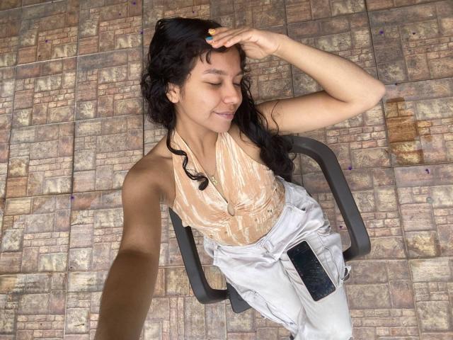 JuliaBennett with wavy black hair, tan halter top, white pants, and black phone, smiling, eyes closed, hand on forehead, taking a selfie from above.