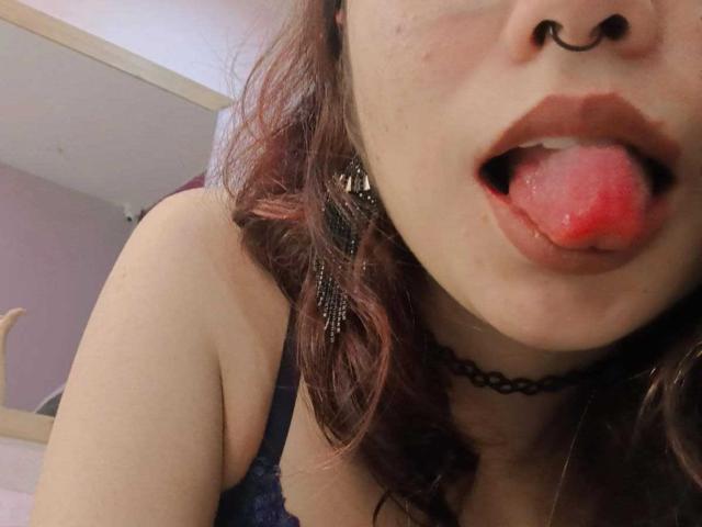 AliCipher with red-tipped tongue sticking out, nose ring, black choker, and dark blue lace top, showing her slightly freckled face and dark brown wavy hair.
