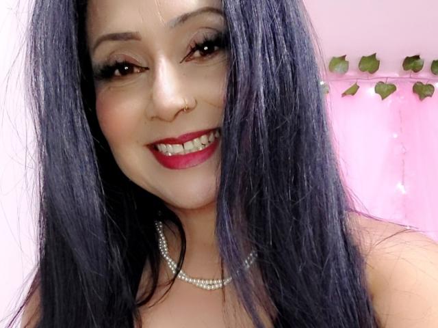 Zaria69 with long black hair, red lipstick, nose ring, and pearl necklace, smiling at the camera. Boobs are not visible. She's wearing a sleeveless top.