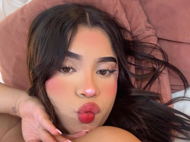 LunaVali with pouty red lips, dark brown eyes, and long black hair, lying on a pink pillow, her hand near her face, showing a small tattoo on her finger.