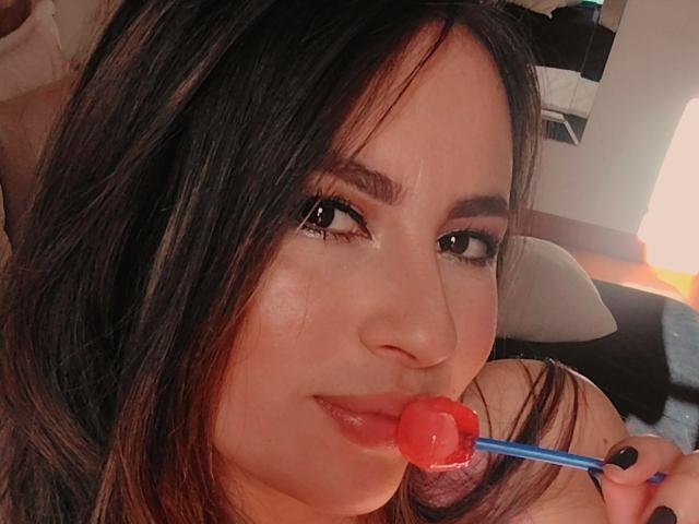 AmiiRosse con capelli scuri, che tiene un lecca-lecca a forma di cuore rosso su un bastoncino blu, sorridendo leggermente, con smalto nero sulle unghie, illuminazione soffusa e trucco leggero..
